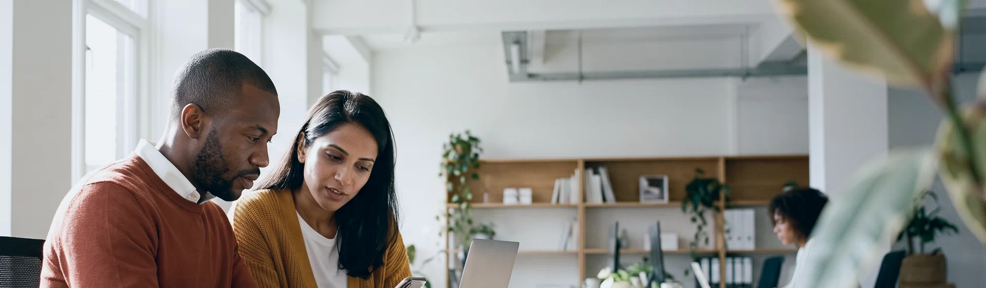 A product lead trying an app on his phone beside a laptop while a colleague leans in to watch, at a sunlit table in an open-plan office.